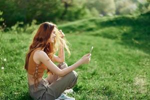 un joven contento mujer sentado en el parque en el verde césped en naturaleza con su teléfono en su manos en su casual ropa y chateando vía vídeo enlace, el concepto de sano recreación meditación foto