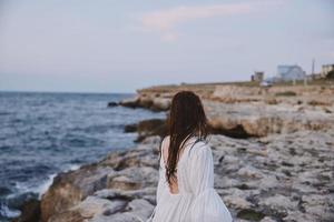 Woman in white dress walks on the beach stones back view photo