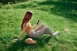 Lanza libre estudiante trabajando en el teléfono sentado al aire libre en el verano en verde césped, móvil y Internet fuera de pueblo foto