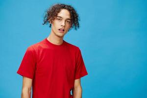 Attractive cute young curly tanned Caucasian guy in basic red t-shirt looks at camera posing isolated on over blue background. Lifestyle and Emotions concept. Good offer with copy space for ad photo