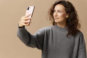 Shocked irritated curly lady in gray casual sweater do video call with phone get bad news posing isolated on over beige background. Social media, network, distance communication concept. Copy space photo