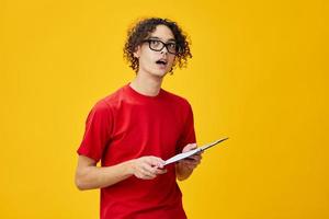 Confused myopic young student man in red t-shirt funny eyewear holds tablet folder with study notes looks aside posing isolated on over yellow background. Free place for ad. Education College concept photo