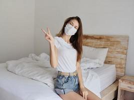 beautiful woman in a medical mask sits on a bed in a bright room and hand gesture positively photo