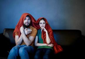 a woman and a man under a red blanket on the couch watching tv in the evening photo