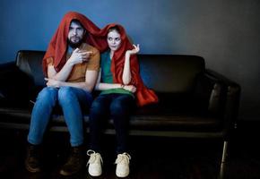 a woman and a man under a red blanket on the couch watching tv in the evening photo