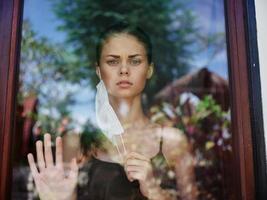 un mujer mira tristemente fuera el ventana en un encierro médico máscara foto