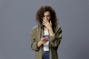 Upset shocked confused curly beautiful woman in casual khaki green shirt receiving bad news using phone posing isolated on over gray blue background. Social Media Influencer concept. Copy space photo
