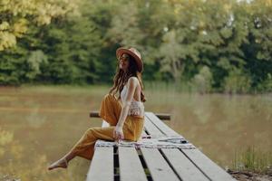 un hippie mujer se sienta en un puente por el río y disfruta el hermosa paisaje alrededor su, contento en el puesta de sol de el otoño Dom foto