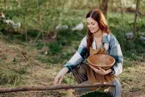 un mujer granjero sonrisas y vierte comida para el aves dentro un pájaro alimentador desde un cuenco a un al aire libre granja a aumento saludable, orgánico pollos foto
