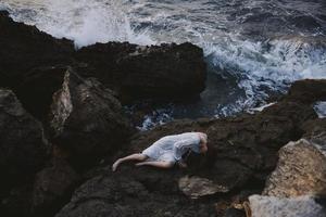 Barefoot woman in long white dress wet hair lying on a rocky cliff view from above photo