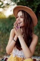 A woman in eco-friendly clothing in a hippie look sits on a colored plaid smiling and looking at an autumn sunset in nature in the park photo