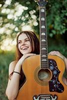 joven hippie mujer con eco imagen sonriente y mirando dentro el cámara con guitarra en mano en naturaleza en un viaje foto