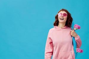 Funny joyful pretty redhead lady in pink hoodie sunglasses with penny board smiling posing isolated on blue studio background. Copy space Banner Offer. Fashion Cinema. Holiday activity photo