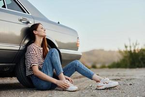 A young woman sits on the ground near her car on the side of the road and looks at the sunset. Camping after a hard road trip photo