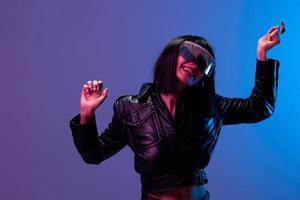 Smiling pretty awesome brunet woman in leather jacket trendy specular sunglasses dance raise hands up posing isolated in blue violet color light background. Neon party Cyberpunk concept. Copy space photo