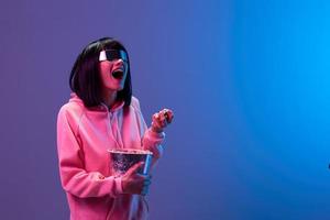 Laughing excited awesome brunet woman in pink hoodie trendy specular sunglasses eat popcorn look aside posing isolated in blue violet color light background. Neon party Cinema concept. Copy space photo