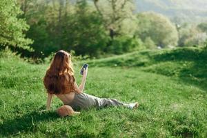 Top view of a woman in an orange top and green pants sitting on the summer green grass with her back to the camera with her phone, a young freelance student's concept of work and leisure photo