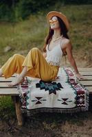 un contento niña sentado en eco ropa en naturaleza en un tartán por el lago vistiendo un sombrero en un hippie Mira y sonriente en preguntarse. otoño estilo de vida foto