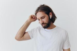 un hombre con un barba y largo pelo en un blanco camiseta y azul pantalones soportes en contra un blanco muro, propensión en contra eso y escuchando a música con inalámbrico blanco auriculares, curioso pensativamente foto