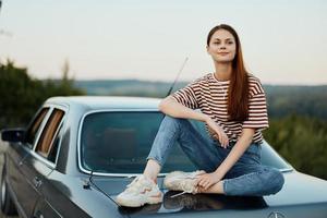 A young woman travels alone in her car on the roads in the countryside and relaxes sitting on the hood watching the sunset photo