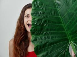 alegre mujer con abierto boca en un traje de baño participación un verde hoja en frente de su exótico de cerca foto