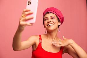 A young woman blogger with colored pink hair and a short haircut takes a picture of herself on the phone and broadcasts a smile in stylish clothes and a hat on a pink background monochrome style photo