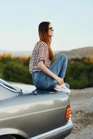 un Moda mujer en elegante anteojos, un a rayas camiseta y pantalones se sienta en el maletero de un coche y mira a el hermosa naturaleza de otoño foto