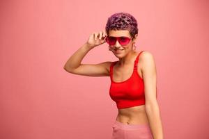 Fashion portrait of a woman with a short haircut in colored sunglasses with unusual accessories with earrings smiling on a pink bright background with a fitness body dancing photo