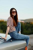 A fashion woman in stylish glasses, a striped t-shirt and jeans sits on the trunk of a car and looks at the beautiful nature of autumn photo