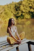 A young happy woman with a hippie smile sits on the lake shore on a bridge and meditates in a lotus pose wearing eco clothing made of natural materials in harmony with nature in the fall photo