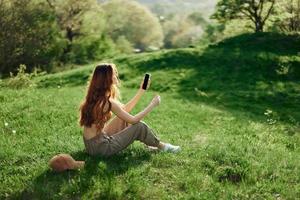 Woman blogger sits on the green grass in a park and takes pictures of herself on her phone against the backdrop of a summer landscape. Young people's lifestyle and concern for the environment photo