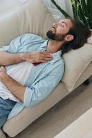 un hombre con un barba duerme en el sofá en el tarde cansado y relajado después estrés y enfermo salud. estrés a trabajar, pobre dormir y salud problemas foto