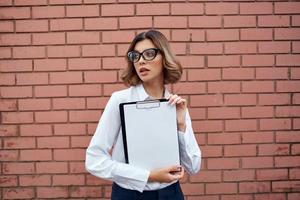 Business woman in white shirt and glasses with documents on the street brick wall photo