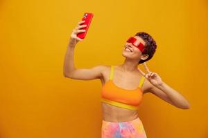 Woman blogger smiling in unusual millennial glasses taking selfies on her phone in sportswear against an orange studio backdrop, free space photo