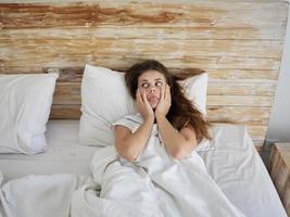 woman lying in bed under the covers holding her face showing tongue photo