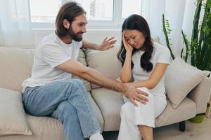 A sad Asian woman talks to a man in tears at home sitting on the couch. Young couple of different nationalities and conflict of interest in a couple photo