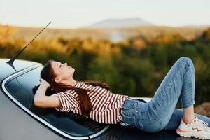 el mujer conductor detenido en el la carretera y laico abajo en el coche a descanso y Mira a el hermosa paisaje en un a rayas camiseta y vaqueros. Complicado viaje a naturaleza foto