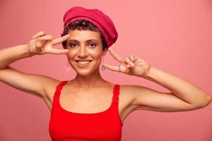 Young athletic woman with a short haircut and purple hair in a red top and a pink hat with an athletic figure smiles and grimaces showing signs with her hands looking at the camera on a pink background photo