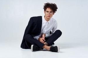 un chico en un camisa, pantalones y zapatillas en el piso en un brillante habitación y un chaqueta en su hombro foto