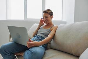 Teenage girl freelancer with laptop sitting on couch at home smiling in home clothes and glasses with short haircut, lifestyle with no filters, free copy space photo