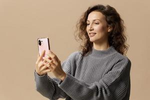 Joyful curly beautiful female in gray casual sweater doing selfie with phone posing isolated on over beige pastel background. Social media, network, distance communication concept. Copy space photo