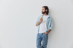 Lanza libre milenario hombre con un barba Bebiendo café desde un reciclado taza en elegante hipster ropa blanco camiseta azul pantalones y camisa en un blanco antecedentes foto