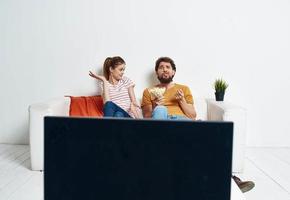 A man and a woman are sitting on the couch in front of the TV and a green flower in a pot indoors photo