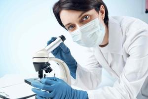 laboratorio asistente vistiendo un médico máscara mirando mediante un microscopio profesional investigación experimentar foto