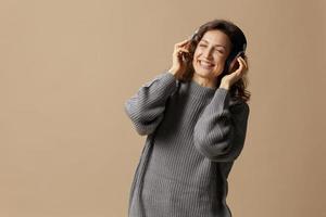 Joyful smiling curly beautiful female in gray casual sweater wear headphones enjoy cool playlist posing isolated on over beige pastel background. Sound streaming platform concept. Copy space photo