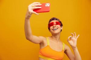 Woman smiles and shows her tongue in unusual glasses millennial takes selfies on her phone in sportswear against an orange studio backdrop, free space photo