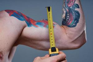 guy measures arm muscles with centimeters on a gray background and a multicolored tattoo bodybuilder fitness photo