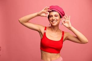 joven atlético mujer con un corto Corte de pelo y púrpura pelo en un rojo parte superior y un rosado sombrero con un atlético figura sonrisas y muecas mirando a el cámara en un rosado antecedentes foto