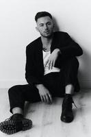 Modelling snapshots. Serious self-confident attractive handsome man in black jacket sitting on floor posing in white studio background. Black and White concept. Copy space photo