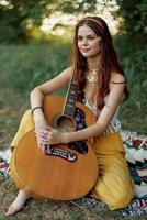 Hippie eco friendly woman with a guitar relaxing in nature sitting on a plaid by the lake in the evening in the rays of the setting sun. A lifestyle in harmony with the body and nature photo